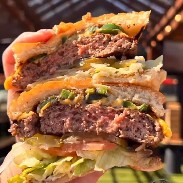 A hand holds a large, juicy burger cut in half, revealing two patties, cheese, jalapeños, and fresh vegetables.