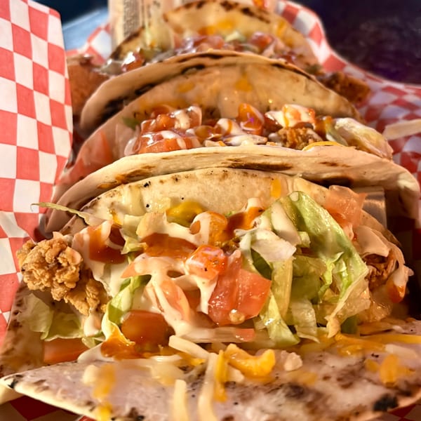 Three chicken tacos with lettuce, tomatoes, cheese, and sauce in a red and white checkered basket.