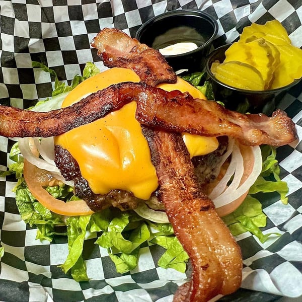 A bacon cheeseburger with lettuce, tomato, and onion, served with pickles and sauce on checkered paper.