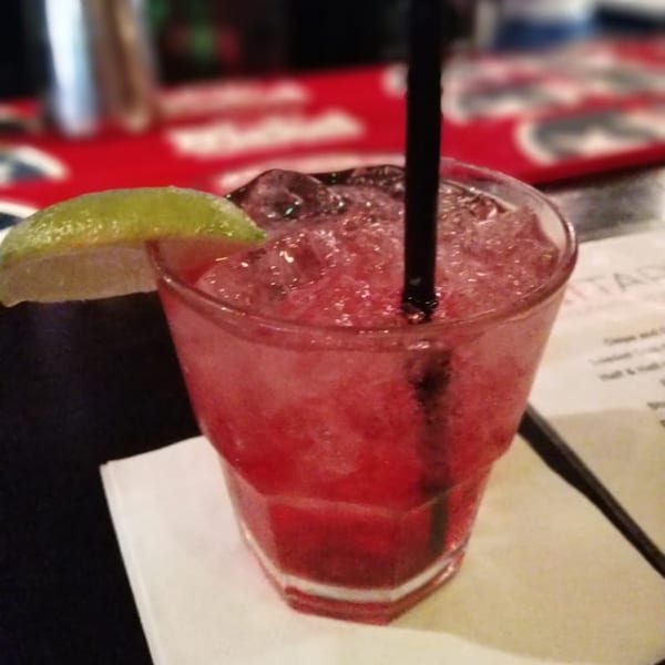 A vibrant red cocktail with ice, a lime wedge, and a black straw, on a bar.