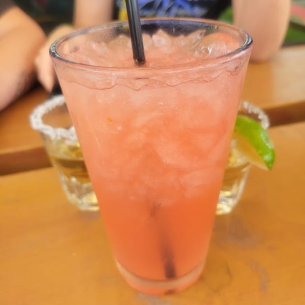 A vibrant pink cocktail with ice and a black straw, next to a shot glass with a lime.